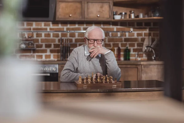 Réfléchi beau homme aux cheveux gris penser et regarder l'échiquier à la maison — Photo de stock