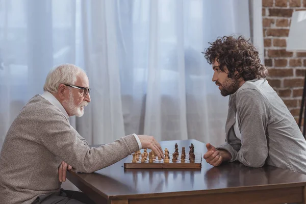 Vista laterale del figlio adulto e padre anziano che gioca a scacchi a casa — Foto stock