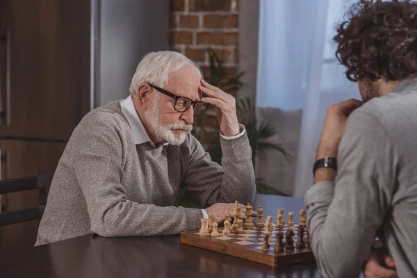 Vue latérale de fils adulte et père aîné pensant tout en jouant aux échecs à la maison — Photo de stock