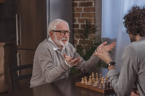 Hijo adulto y padre mayor haciendo gestos mientras juega al ajedrez en casa - foto de stock