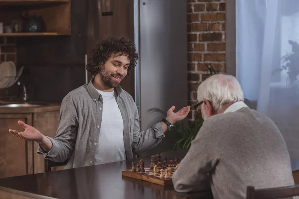 Hijo adulto haciendo gestos mientras juega al ajedrez con padre mayor en casa - foto de stock