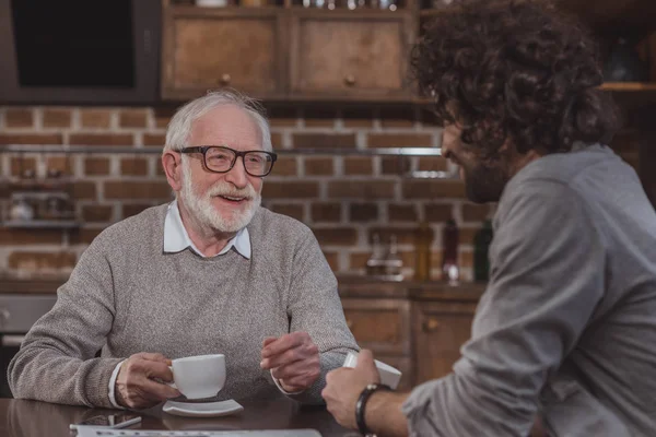 Fils adulte et père aîné parler et boire du café à la maison — Photo de stock