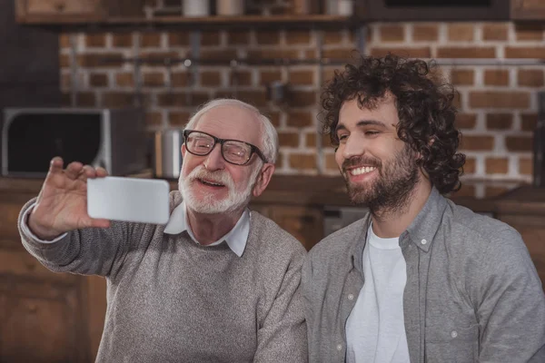 Sonriente hijo adulto y padre mayor tomando selfie en cocina - foto de stock