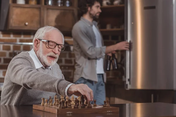 Hijo adulto mirando en nevera y padre mayor jugando ajedrez en casa - foto de stock
