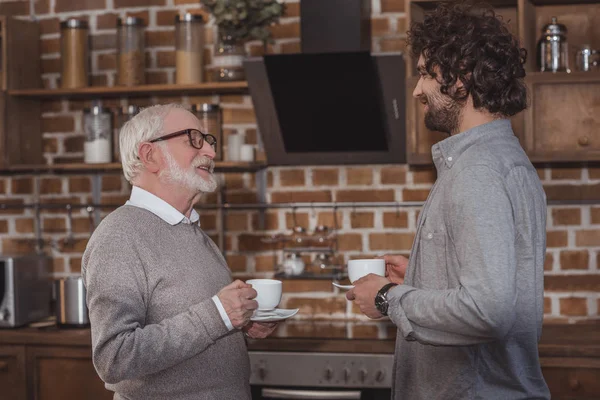 Взрослый сын и старший отец стоят с чашками кофе на кухне — стоковое фото