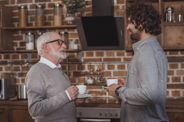 Fils adulte et père aîné debout avec des tasses de café dans la cuisine — Photo de stock