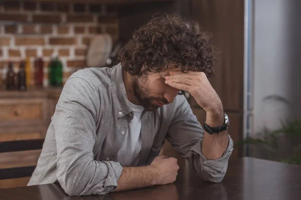 Hombre cansado sentado en la mesa en la cocina y tocando la cabeza - foto de stock