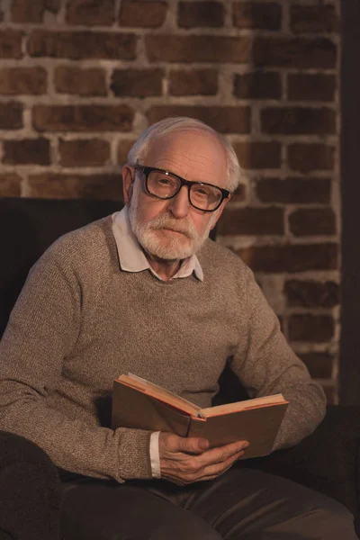 Handsome grey hair man holding book and looking at camera at home — Stock Photo