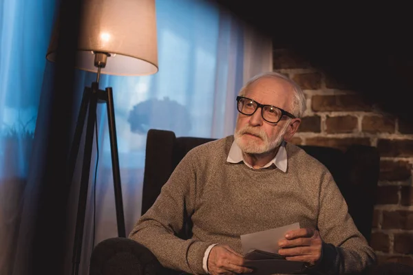 Handsome grey hair man sitting in armchair, holding old photos and looking away — Stock Photo