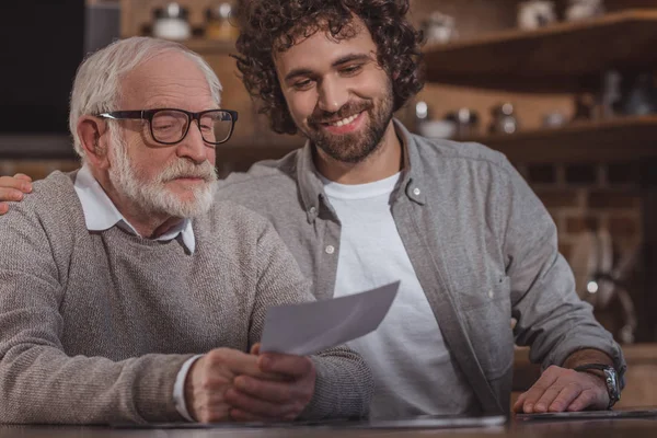 Sourire fils adulte étreignant père aîné et regardant des photos à la maison — Photo de stock