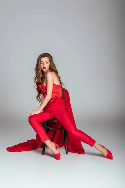 Belle femme à la mode en vêtements rouges assis sur chaise, sur gris — Photo de stock