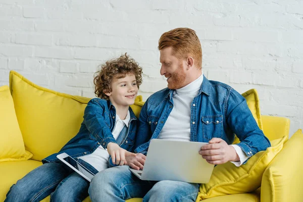 Pai e filho sentados no sofá com laptop e tablet — Fotografia de Stock