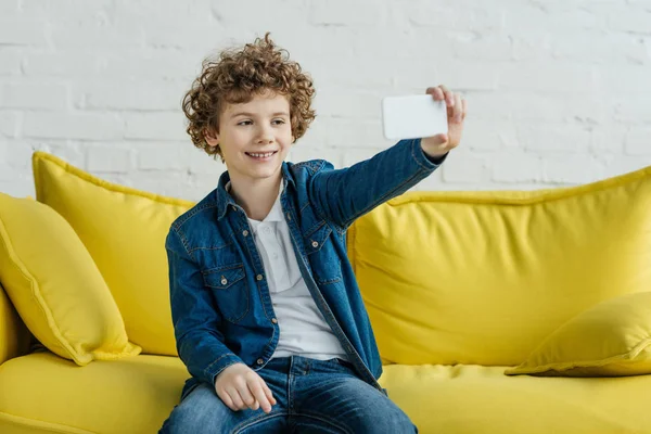 Niño sonriente tomando selfie sentado en el sofá - foto de stock