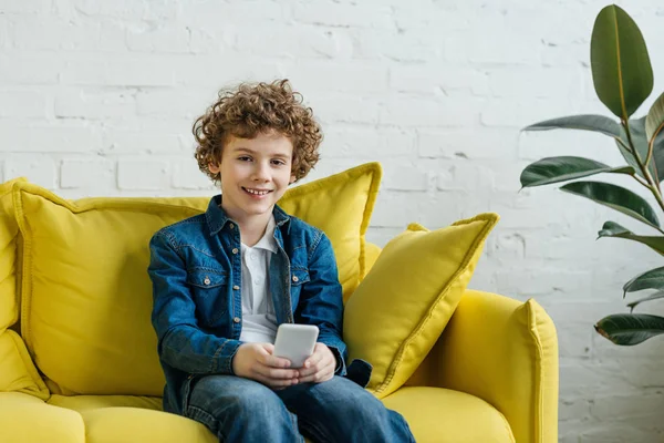 Sonriente niño usando el teléfono sentado en el sofá - foto de stock