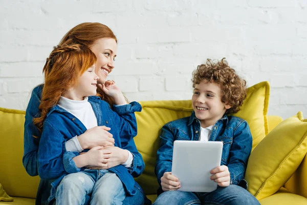 Abrazando a la madre y a la hija sentadas en el sofá con el hijo sosteniendo la tableta - foto de stock