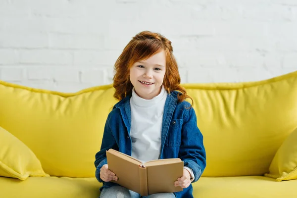Sonriente niño pequeño sosteniendo libro en el sofá - foto de stock