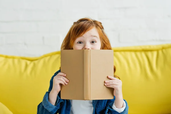 Child sitting on sofa and holding book in front of her face — Stock Photo