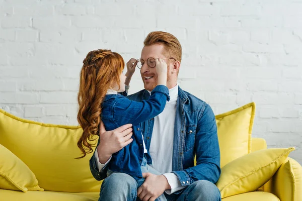 Hija y padre engañando con gafas mientras está sentado en el sofá - foto de stock