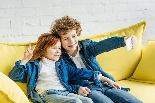 Усміхнені діти беруть селфі, сидячи на дивані — стокове фото