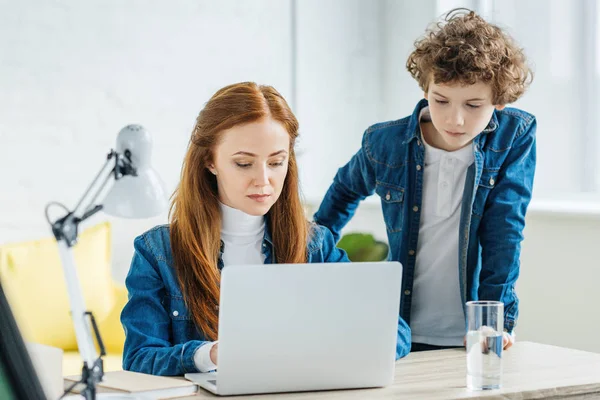 Enfant debout par une femme travaillant avec un ordinateur portable à la table — Photo de stock