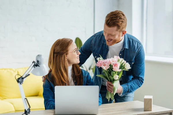 Homme surprenant jeune femme travaillant par ordinateur portable avec un bouquet de tulipes — Photo de stock