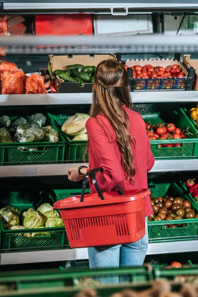 Vista posterior de la mujer con cesta de la compra elegir verduras frescas crudas en la tienda de comestibles - foto de stock