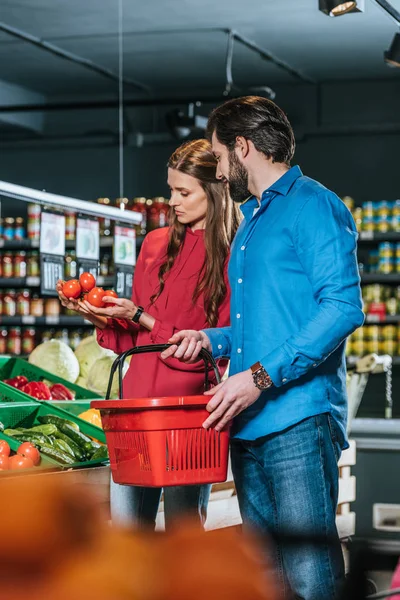 Pareja con cesta de la compra elegir verduras frescas juntos en el supermercado - foto de stock