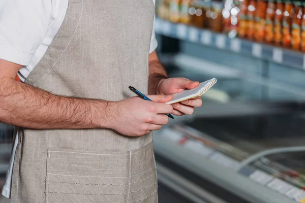 Partial view of shop assistant in apron with notebook in supermarket — Stock Photo
