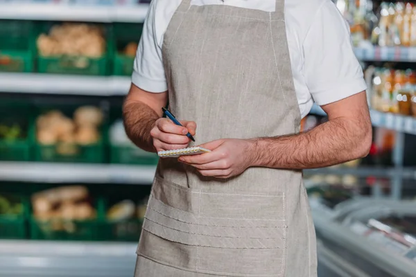 Teilansicht einer Verkäuferin in Schürze, die im Supermarkt Motten in Notizbuch bastelt — Stockfoto