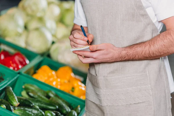 Teilansicht einer Verkäuferin in Schürze, die im Supermarkt Motten in Notizbuch bastelt — Stockfoto