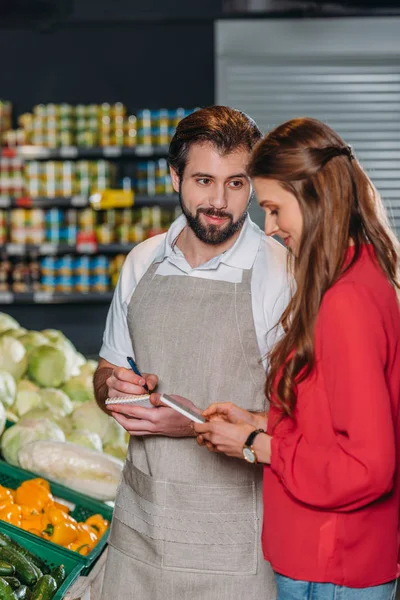Asistente de tienda con notebook y comprador femenino en el supermercado - foto de stock