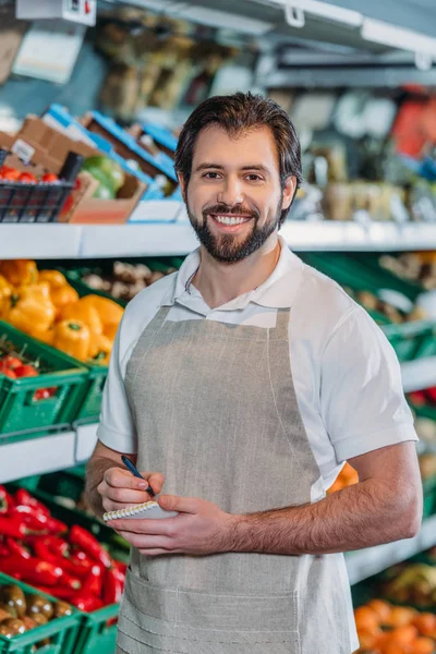 Ritratto di commessa sorridente in grembiule con taccuino nel supermercato — Foto stock