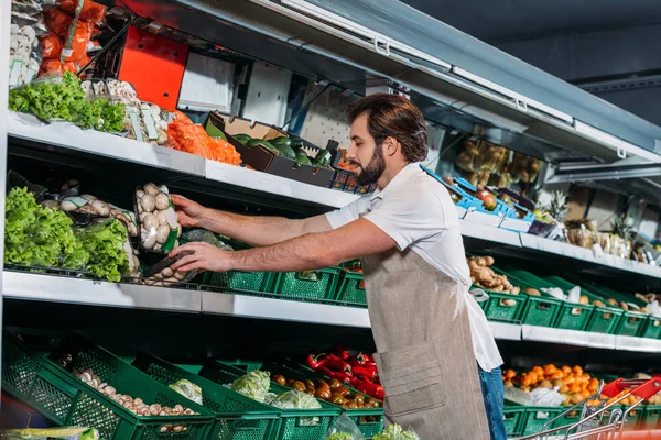 Мужчина помощник в фартуке организация свежих овощей в продуктовом магазине — стоковое фото