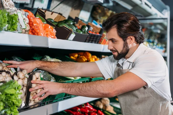 Asistente de tienda que organiza verduras frescas en la tienda de comestibles - foto de stock