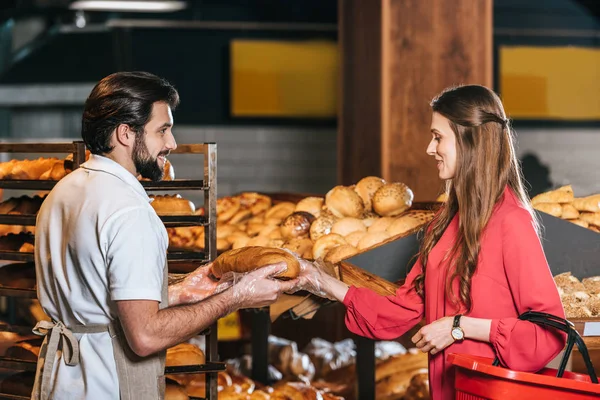 Side view of shop assistant giving loaf of bread to woman with shopping basket in supermarket — Stock Photo
