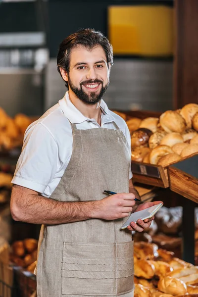 Portrait of smiling male shop assistant in apron with notebook in supermarket — Stock Photo