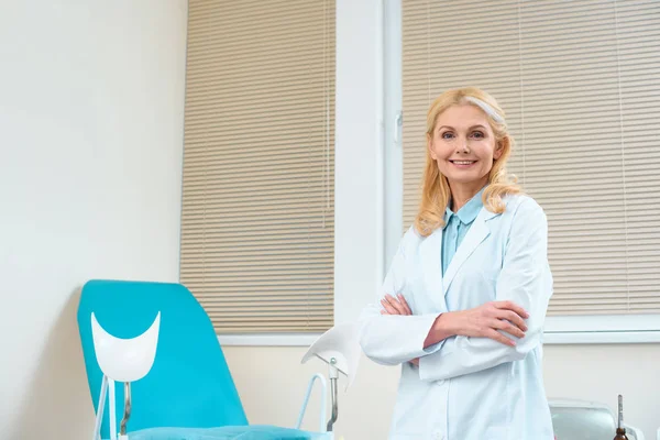 Sorridente ginecologa femminile con braccia incrociate davanti alla sedia ginecologica — Foto stock