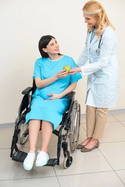 Mature obstetrician gynecologist giving green apple to pregnant woman on wheelchair — Stock Photo
