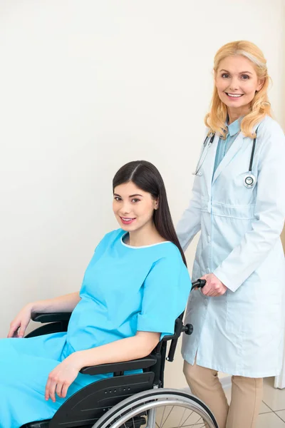 Happy obstetrician gynecologist riding pregnant woman on wheelchair at maternity hospital and looking at camera — Stock Photo