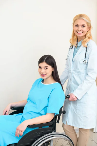 Ginecólogo obstetra feliz montar mujer embarazada en silla de ruedas en el hospital de maternidad y mirando a la cámara - foto de stock