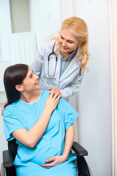 Happy obstetrician gynecologist riding pregnant woman on wheelchair at maternity hospital and supporting her — Stock Photo