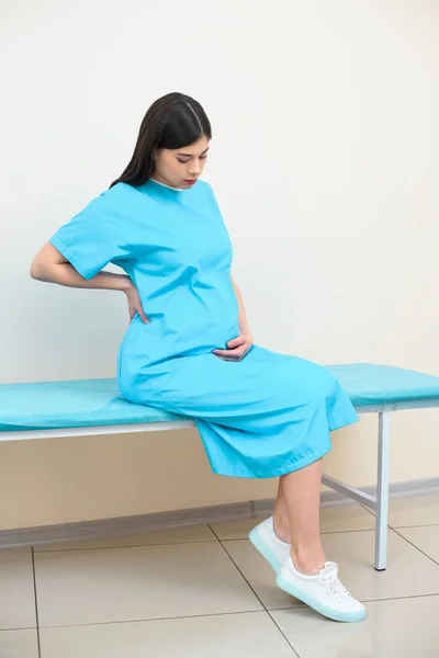 Joven embarazada sentada en la cama en el hospital de maternidad y reteniéndola - foto de stock