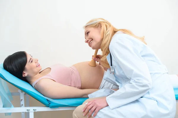 Акушер-гінеколог, який слухає серцебиття дитячого плоду щасливої вагітної жінки зі стетоскопом Траубе — стокове фото