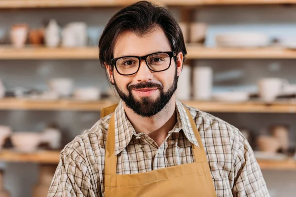 Бородатый гончар в фартуке и очки в мастерской — стоковое фото