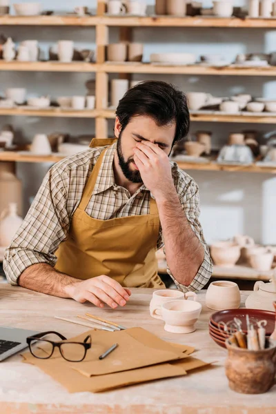 Alfarero macho cansado en delantal sentado a la mesa con cerámica en el taller - foto de stock