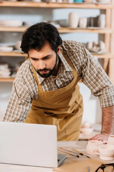 Homme dans tablier travaillant avec ordinateur portable en atelier de poterie — Photo de stock