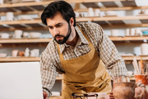 Male potter in apron working with laptop in workshop — Stock Photo