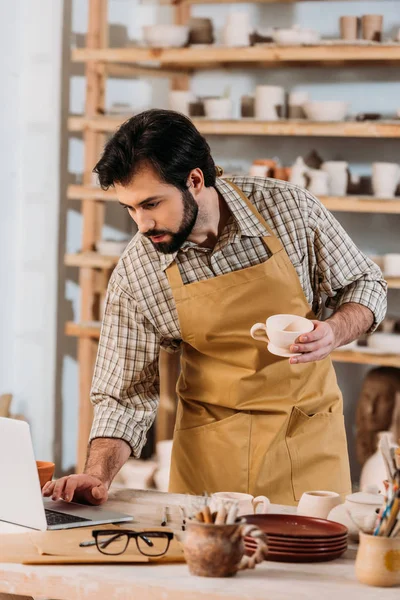 Male potter in apron holding ceramic cup and using laptop in workshop — Stock Photo