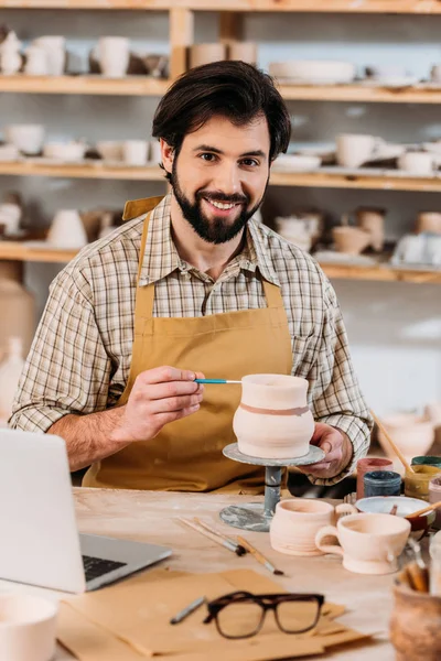 Oleiro macho feliz pintura cerâmica louça na oficina com laptop na mesa — Fotografia de Stock