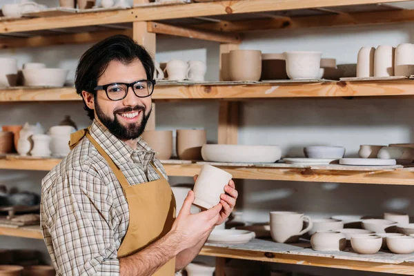 Alfarero sonriente sosteniendo taza de cerámica en el taller con vajilla en los estantes - foto de stock